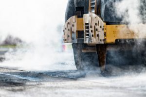 A close-up of a vibrating asphalt compactor (steam roller) rolling over freshly laid asphalt during the resurfacing of a rural highway. The road surface was milled of the old top layer of asphalt and that material is being recycled into aggregate for new asphalt